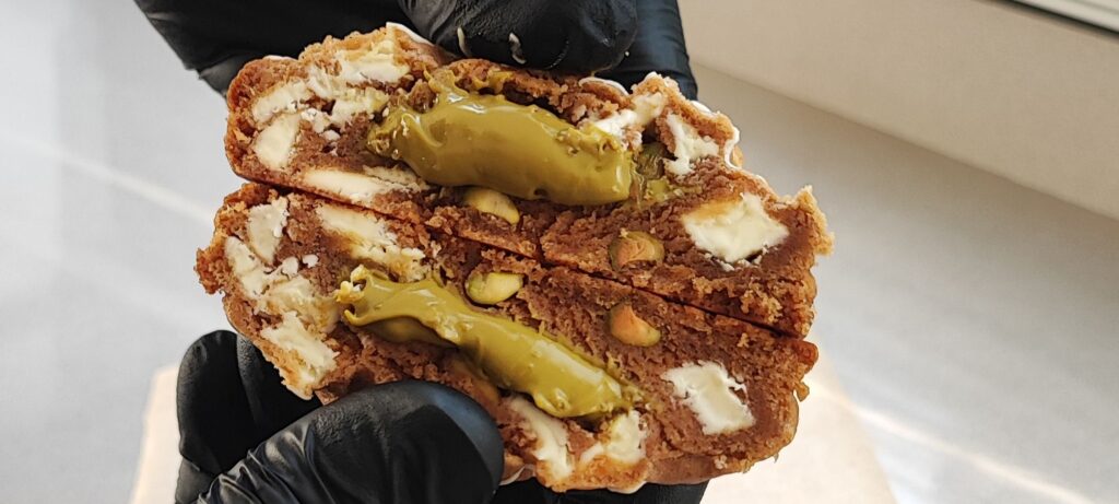 Primer plano de la cookie Pistacho Kiss de Sweet Cocoa sostenida entre las manos, cookie artesanal estilo New York con pistacho.
